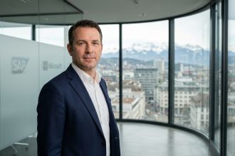 Consultant IT senior de TVH Consulting en blazer marine devant une baie vitrée avec vue sur les Alpes suisses et un skyline urbain moderne
